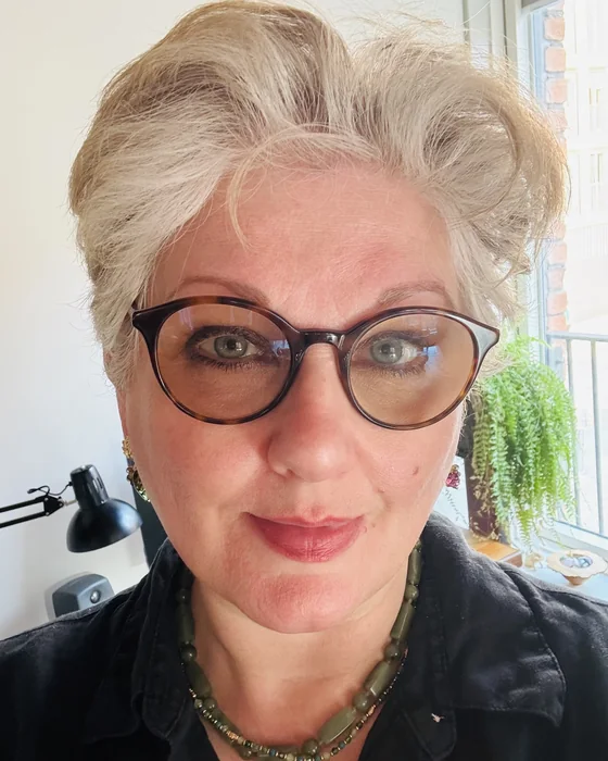 Emma Clarke, smiling, in round tortoiseshell glasses, at her desk by a window in Manchester.